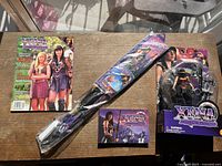 Photo showing all items in the lot placed on a wooden table: the packaged Xena action figure, a magazine, a folded poster, and a booklet.