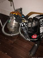Dyson vacuum unit with clear dirt canister, orange cyclone top, hose, and some attachments, photographed on dark floor next to bags.