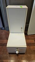 Tall narrow white metal locked drawer with key inserted at front, wear on finish visible.