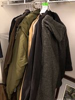 Seven men’s jackets hanging on rack showing various colors and materials including quilted green and fleece gray with hood.