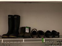 View showing two Griffin tall black rubber boots, brown lace-up boots, and three pairs of Crocs on shelf.