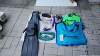 Wide shot showing all snorkeling gear: fins, mask, snorkel, green mesh bag with accessories, and two floatation vests (green and blue) labeled 'The Floater by Snorkel Bob'. Items on tiled floor showing wear.