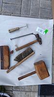 Photo showing the six tools laid out including two wooden mallets, the brush, the wood plane, the spike, and the masonry tool. All items are on a neutral background for clear viewing.