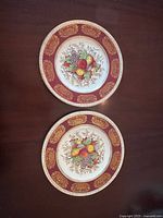 Two antique china plates placed on a wooden table showing the fruit design and gold-accented maroon decorative border.