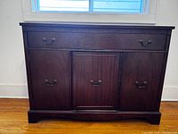 Front view of antique mahogany china cabinet showing three doors and two drawers with brass handles, flush against a wall below a window.