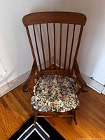 Front view of the antique wooden rocking chair with floral cushion on the seat, showing vertical spindle back and curved armrests.