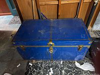 Front view of blue metal antique trunk with corner reinforcements, locks, and keyhole, set on marble floor.