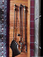 Fire tools laid out on floor showing poker, tongs, shovel with shell design, and small brush with hanging chains, all with brass handles.