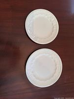 Two white scalloped edge plates stacked on a wooden surface, showing the front decorative side.