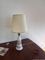 Photo of a white ceramic table lamp with brown decorative accents and a white fabric shade, lying on a wooden surface.