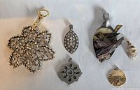 Six assorted pendants laid flat on a white surface, showing various designs and materials including metal, rhinestones, wood, enamel, and resin.