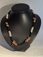 Front view of black seed bead twisted necklace with clusters of faux pearls, orange coral beads, and green glass beads displayed on black bust.