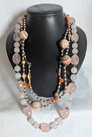 Photo of three layered beaded necklaces displayed on a black jewelry bust, showing variety of bead shapes and colors including rose quartz and ceramic.