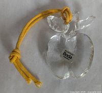 Front view showing clear glass apple-shaped ornament with leaves and stem, with yellow cord loop attached and Boda Sweden sticker on front.