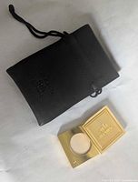 Gold-tone 5th Avenue solid perfume compact open showing the solid perfume inside, with black drawstring pouch next to it.