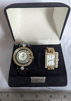 Pair of women's Elizabeth Taylor wristwatches in black watch box, showing round watch with leather strap and rectangular watch with metal bracelet.