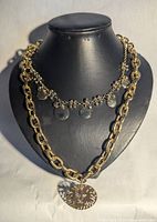 Two necklaces displayed on black bust. Gold-tone chain is chunky with large links and lion face pendant with cutout design. Silver-tone chain is delicate with small round coin-like charms evenly spaced.