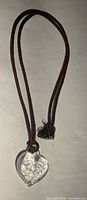Full view of the necklace showing the clear glass heart pendant hanging on a brown fabric cord with knot and tasseled ends.