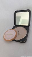 Open black square powder compact showing beige matte powder, applicator puff with Lancôme logo, and built-in mirror.