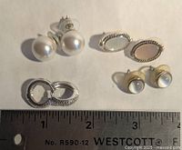 Four pairs of mixed material earrings arranged on a flat surface next to a ruler for scale. Includes pearl studs, metallic oval studs, round studs, and hoop earrings.