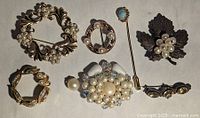 Top view of seven assorted vintage brooches and pins on a white background, showing gold-tone and silver-tone metals, faux pearls, rhinestones, turquoise-tone beads, with floral and leaf decorative designs.