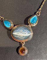 Close-up of the pendant showing central agate-colored cameo with etched pattern, flanked by turquoise teardrop inlays and amber round drop below.