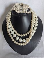 Close-up of three-strand simulated pearl necklace displayed on black bust, showing graduated cream and white faux pearls and decorative clasp.