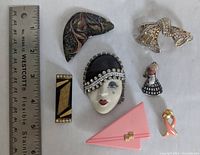 Lot of 8 vintage brooches and pins arranged with a ruler for size reference, showing varied designs and materials including plastic, enamel, metal, and rhinestones.