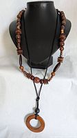 Full view of the long wooden cord necklace with a circular wooden pendant and the beaded medium-length wooden necklace displayed on a black necklace bust.