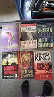 Top view of six paperback books: "Ya-Yas in Bloom," "Summer Sisters," "Dark Summer," "Murder Interrupted," "Highland Champion," and "Cujo." Shows cover condition with some edge wear and scuffs.