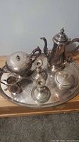 Full view of silver-plated tea and coffee set on oval tray, showing all pieces including teapot, coffee pot, creamer, sugar bowl with lid, waste bowl with lid, and 2 candlesticks.