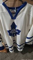 Front view of white hockey jersey with large blue Toronto Maple Leafs logo showing the team's name and leaf motif.