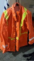 Front view of the orange rain poncho showing snap button front closure, high-visibility reflective stripes on sleeve and across chest, and drawstring hood.