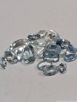 Multiple loose genuine aquamarine gemstones of various cuts and shapes spread on a plain surface.