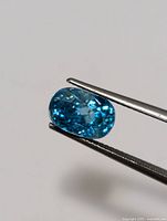 Close-up photo showing the oval cut, bright blue zircon held with tweezers, emphasizing the gemstone's brilliance and facets.