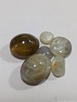 Six polished genuine cat's eye cabochon gemstones displayed on white surface showing natural chatoyancy effect and inclusions.