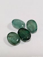 Four oval-shaped faceted green emerald gemstones displayed on white background showing color variations and internal inclusions.