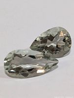 Two pear-shaped light green faceted gemstones lying side by side on a white surface