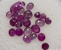 Close-up photo of 22 loose ruby gemstones varying in shape and color laid out on a white surface.