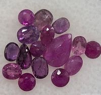 Close-up view of 15 assorted faceted ruby stones in mixed cuts and shades of pink-purple on white background.