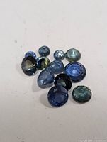 Multiple faceted sapphires of varying sizes and shapes on a white background, displaying different shades of blue and greenish-blue, cut and polished.