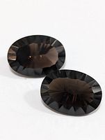 Two oval-shaped dark faceted gemstones with smoky coloration, lying side-by-side on a white background.
