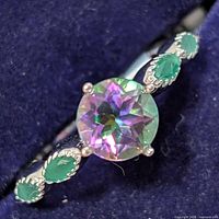 Close-up photo of silver ring with bright iridescent round mystic topaz center stone and three pear-shaped green stones on each side in decorative silver setting, on dark blue background.