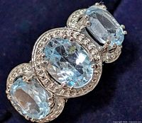 Close-up photo of the silver ring showing the three oval-cut blue topaz stones with silver detailing around them against dark background.