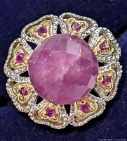 Close-up top view of silver ring with a large central round faceted ruby and surrounding smaller rubies in a decorative setting.