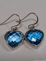 Front angle close-up of the pair of silver earrings with bright blue heart-shaped faceted topaz stones and hook ear wires.