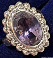 Close-up photo of a sterling silver ring with a large oval faceted amethyst gemstone in the center and a decorative silver setting with bead and rope details.