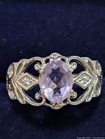 Close-up front view of silver ring with faceted oval amethyst center stone and delicate scrollwork band with two diamond-shaped settings each holding a small clear stone.