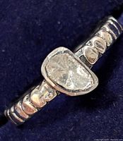 Close-up of silver ring showing asymmetrical diamond and textured band against dark background.