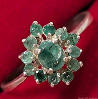 Close-up front view showing central oval emerald surrounded by smaller emerald stones in a floral cluster on a silver band.